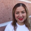 Close-up portrait of a woman smiling with red lipstick against a textured wall.