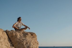 Man in meditation pose on a rocky cliff with ocean view at sunset, embracing serenity and nature.